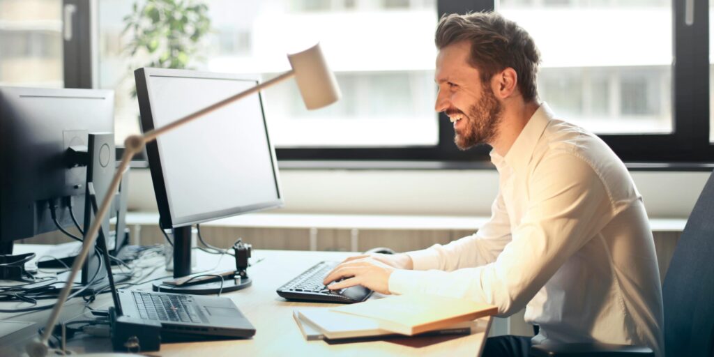 A man smiling while working at an office desk with a computer and natural daylight streaming in through large windows.